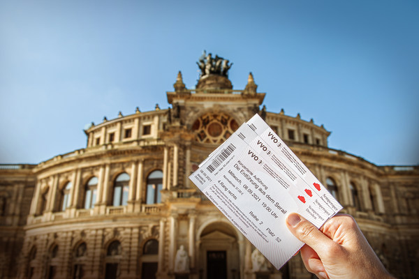 Eine Hand hält Eintrittskarten für die Semperoper in die Höhe, im Hintergrund ist die Semperoper zu sehen