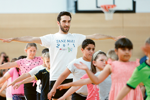 Ein Balletttänzer und mehrere Schülerinnen und Schüler tanzen mit ausgestreckten Armen in einer Turnhalle.